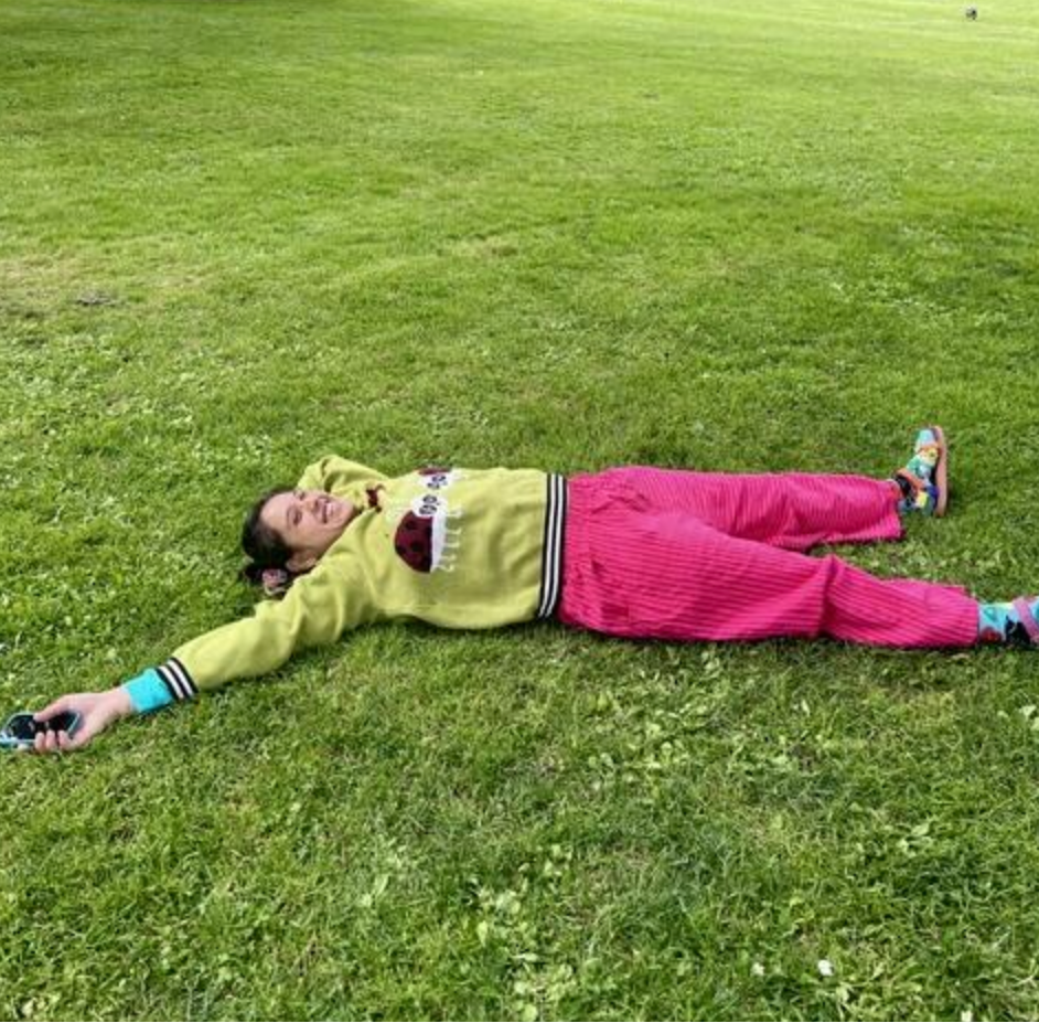 A person lying flat on a green grass lawn in a park, wearing a lime green sweatshirt with black and white stripes on the sleeves and hem, a dark red chest patch, and bright pink pants.