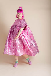 A person with bright pink hair wearing a voluminous, crinkled metallic pink dress, standing against a solid beige studio background.
