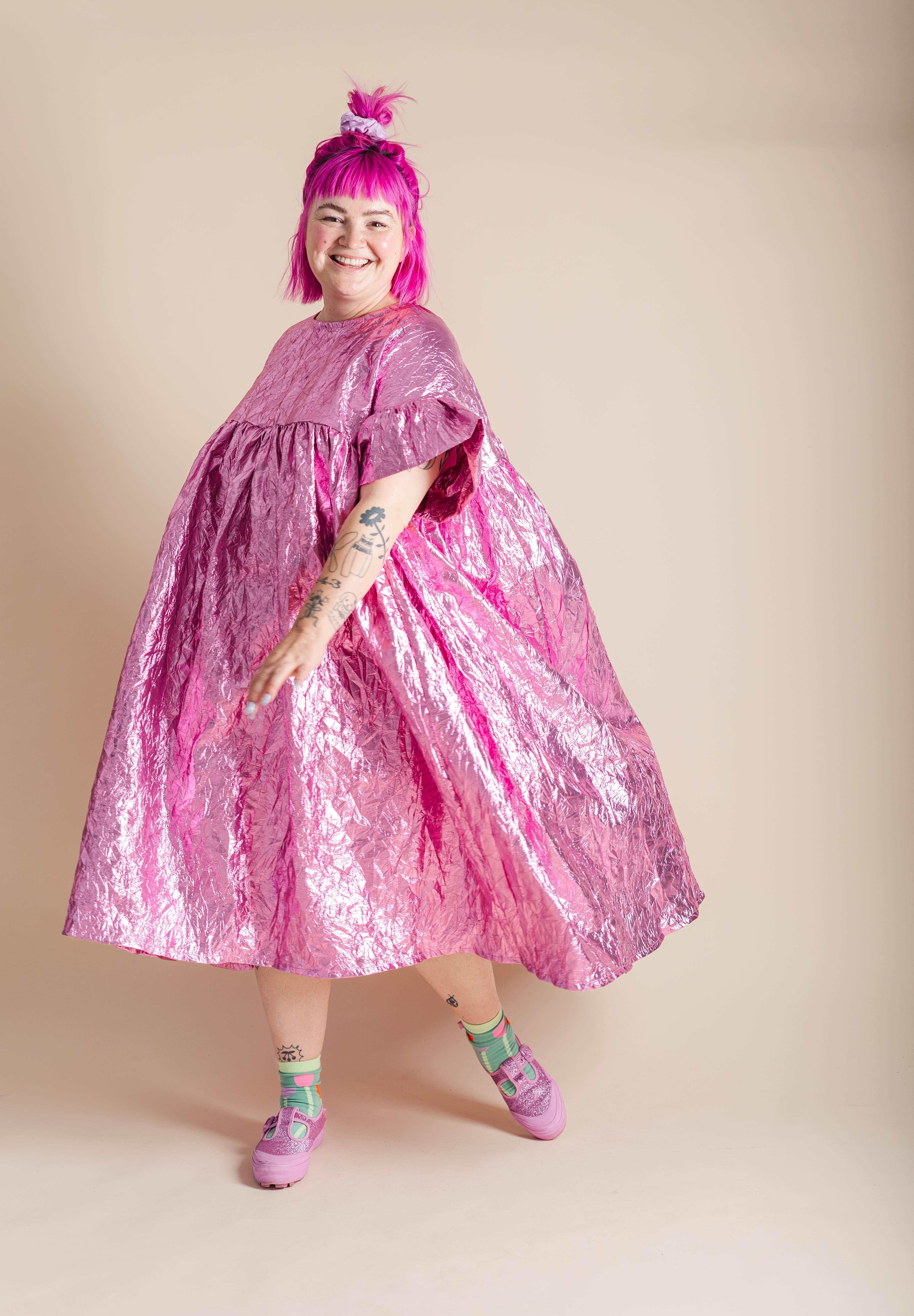 A person with bright pink hair wearing a voluminous, crinkled metallic pink dress, standing against a solid beige studio background.