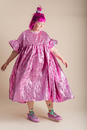 A person with bright pink hair wearing a shiny metallic pink dress, styled with colorful socks and pink sneakers, standing against a solid beige background.