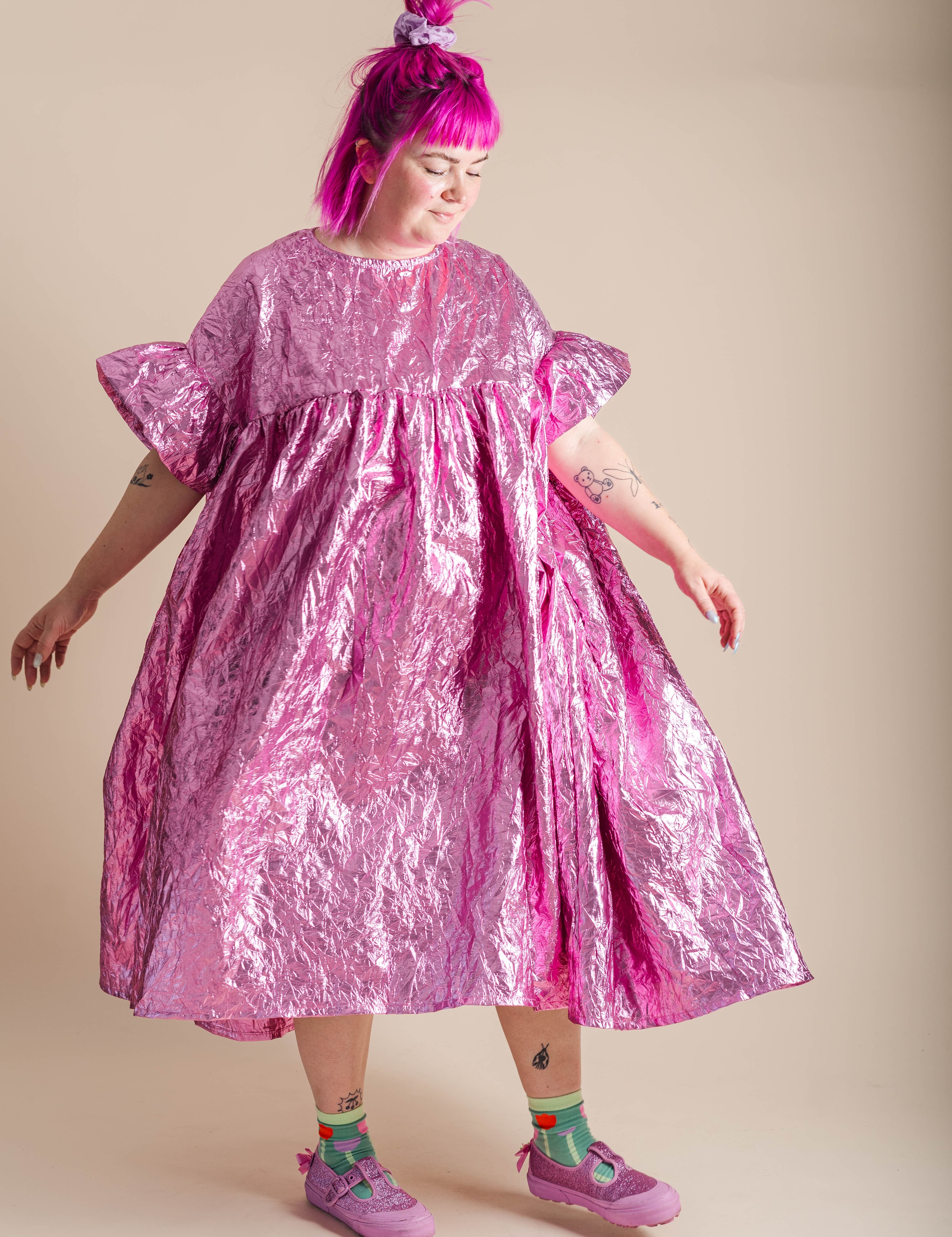 A person with bright pink hair wearing a shiny metallic pink dress, styled with colorful socks and pink sneakers, standing against a solid beige background.