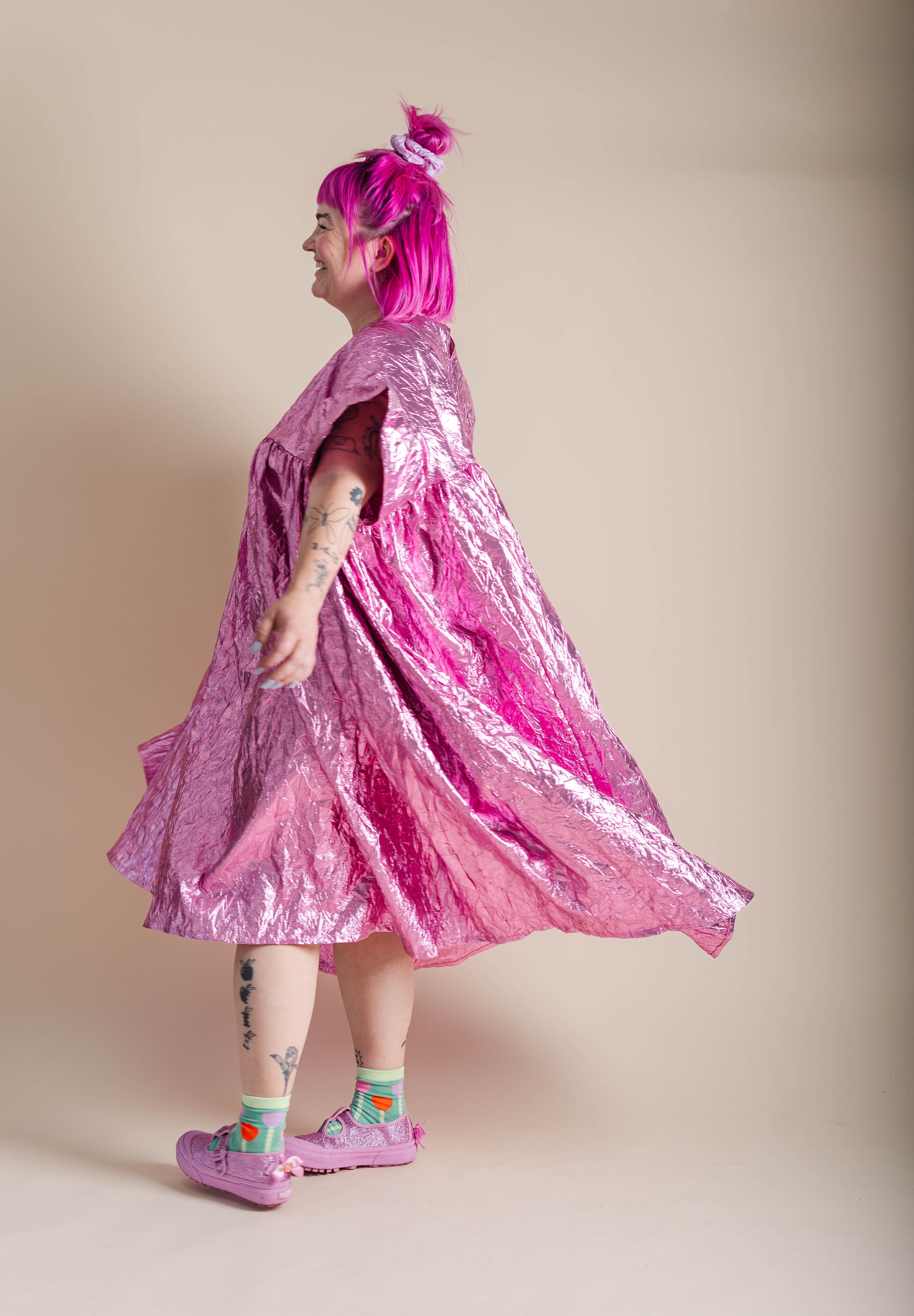 A person with bright pink hair stands in profile wearing a shiny metallic pink cape-like dress or outerwear piece with a voluminous skirt, photographed against a plain beige studio background.
