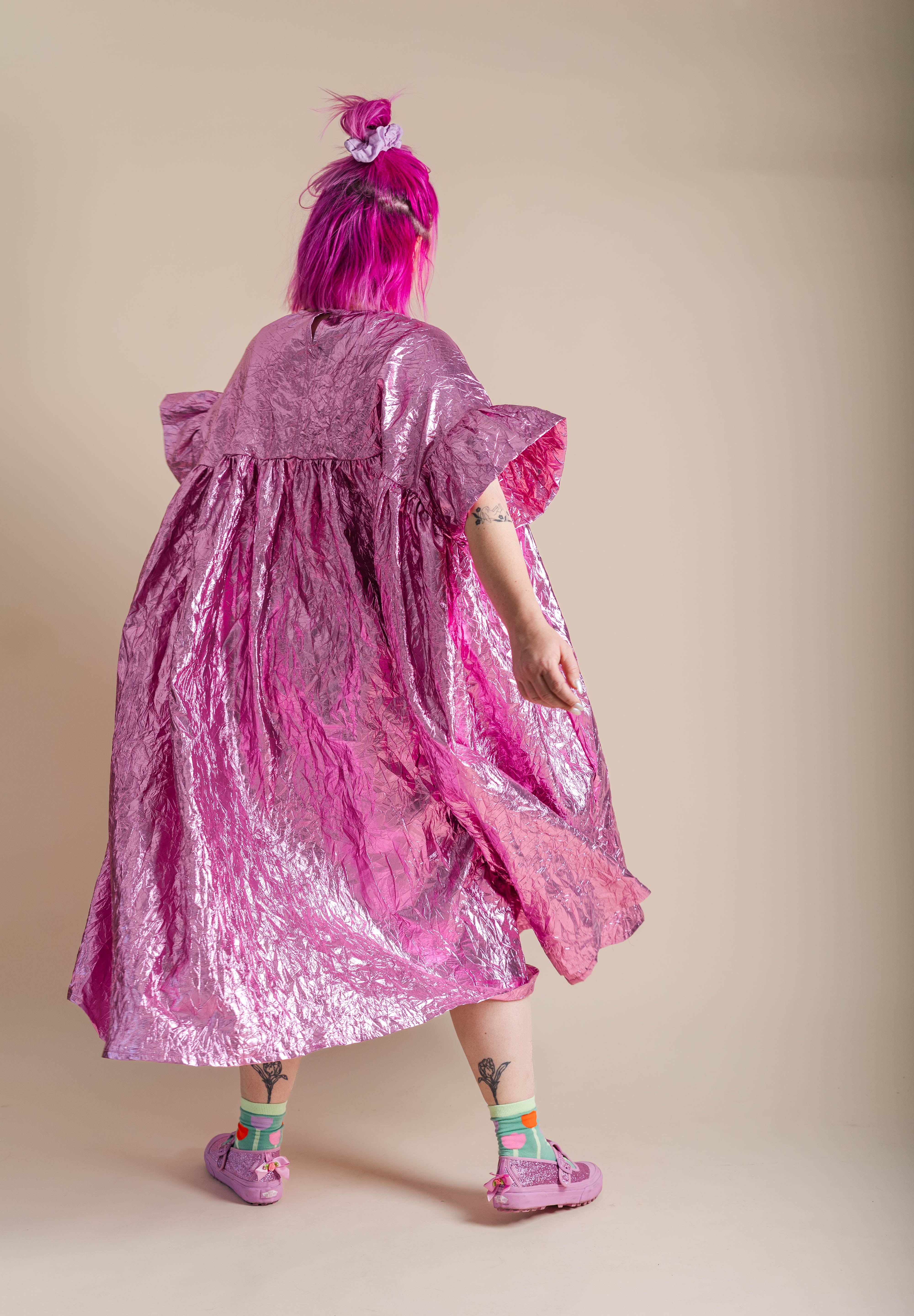 A person with bright pink hair is shown from behind wearing a voluminous, crinkled metallic pink dress and matching pink shoes against a plain beige studio background.