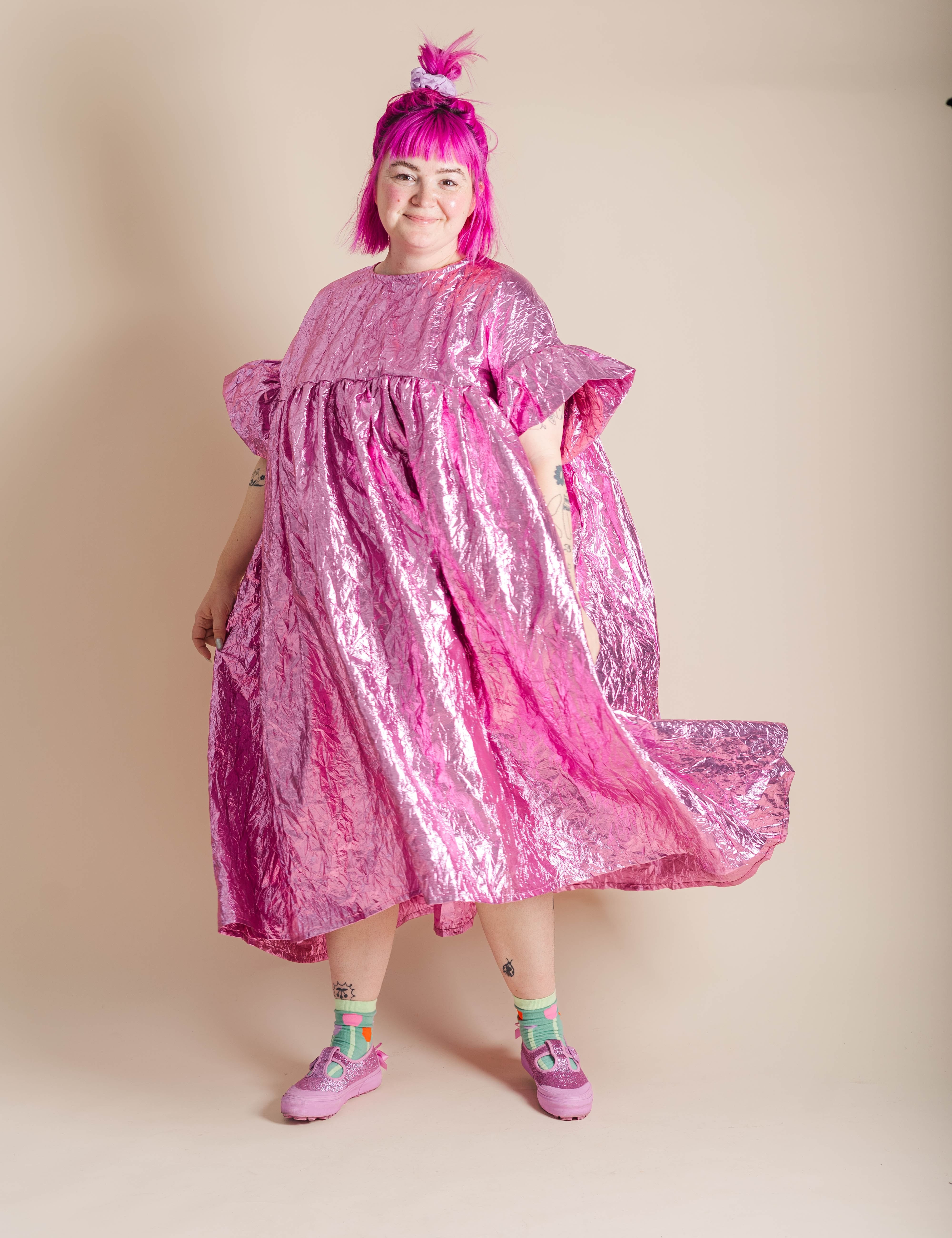 A person with bright pink hair wearing a shiny metallic pink dress and pink shoes, standing against a plain beige studio background.