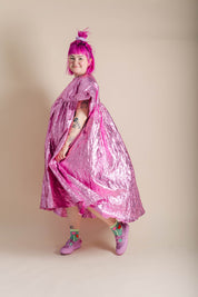 A person with bright pink hair poses in a shiny metallic pink cape-like garment against a plain beige studio background.