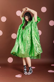 A model wearing a shiny, crinkled bright green poncho-style dress, posing with her arms raised against a brown backdrop decorated with pink polka dots.
