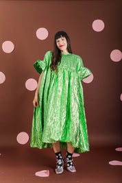 A woman modeling a shiny metallic lime green midi dress with short puff sleeves, standing against a dark brown backdrop decorated with pink polka dots.
