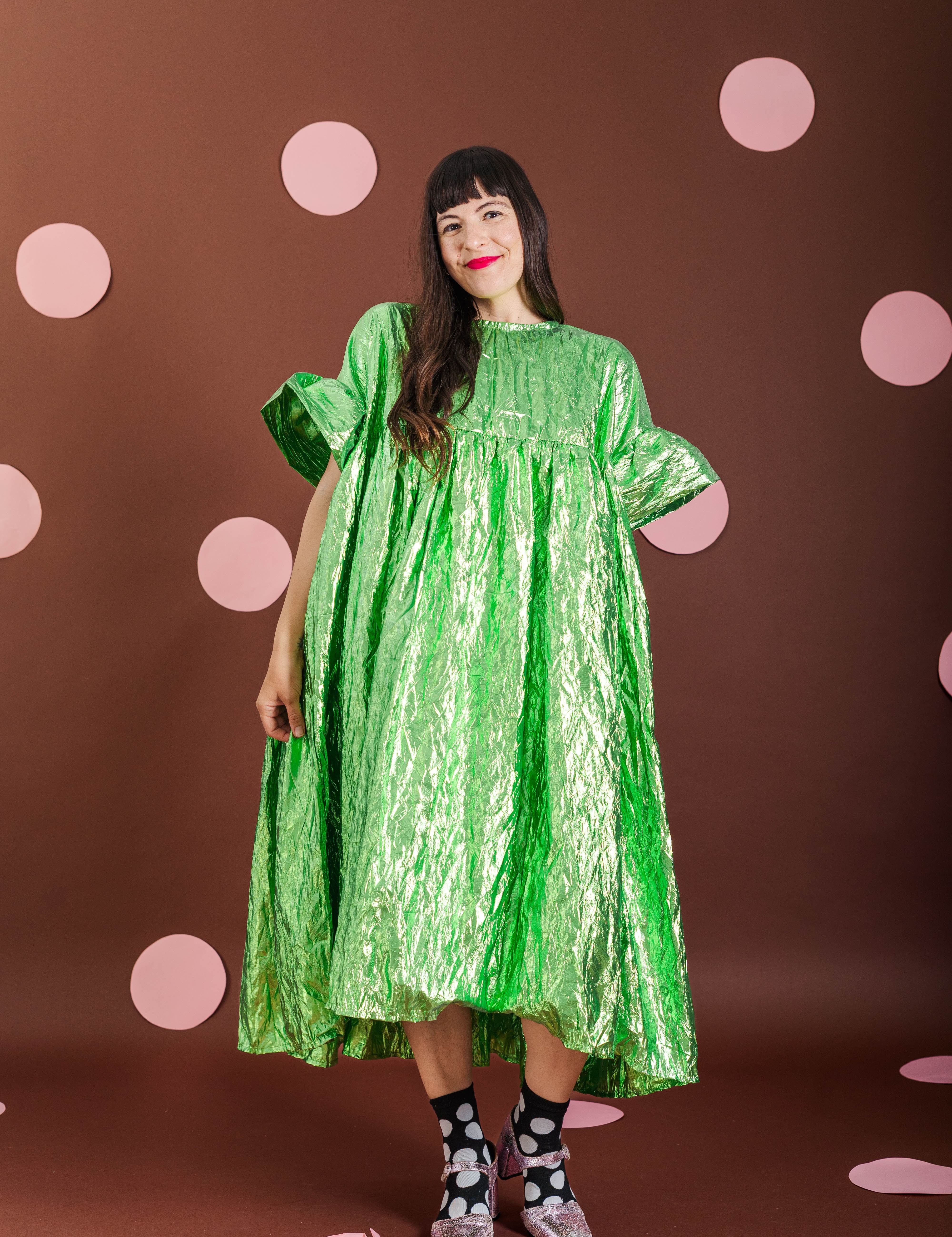 A woman modeling a shiny metallic lime green midi dress with short puff sleeves, standing against a dark brown backdrop decorated with pink polka dots.