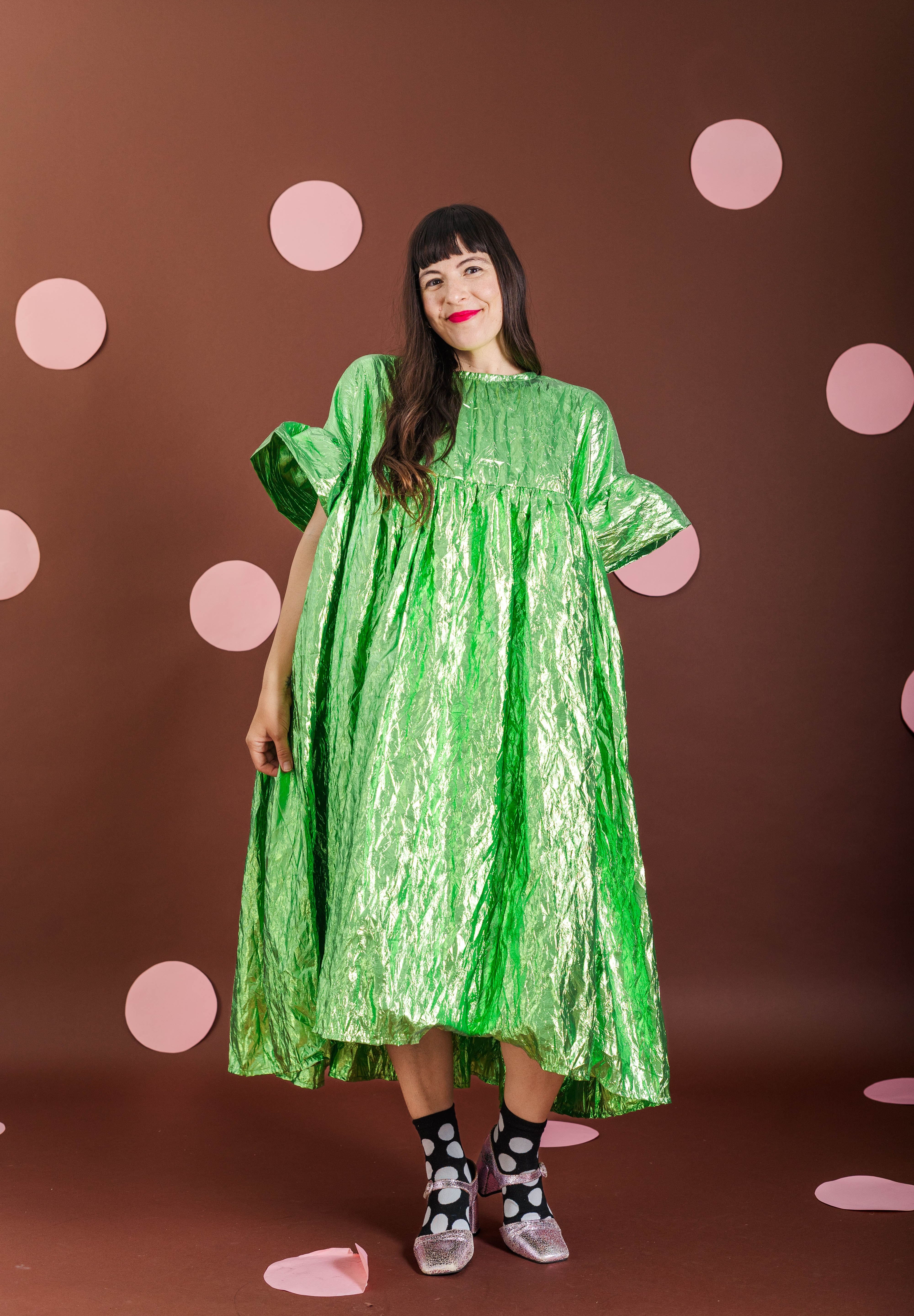 A woman modeling a shiny metallic lime green midi dress with short puff sleeves, standing against a dark brown backdrop decorated with pink polka dots.