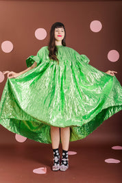 A woman wearing a voluminous, shiny bright green dress with short puff sleeves and a high-low hem, holding the dress out to show its shape against a brown backdrop with pink polka dots.