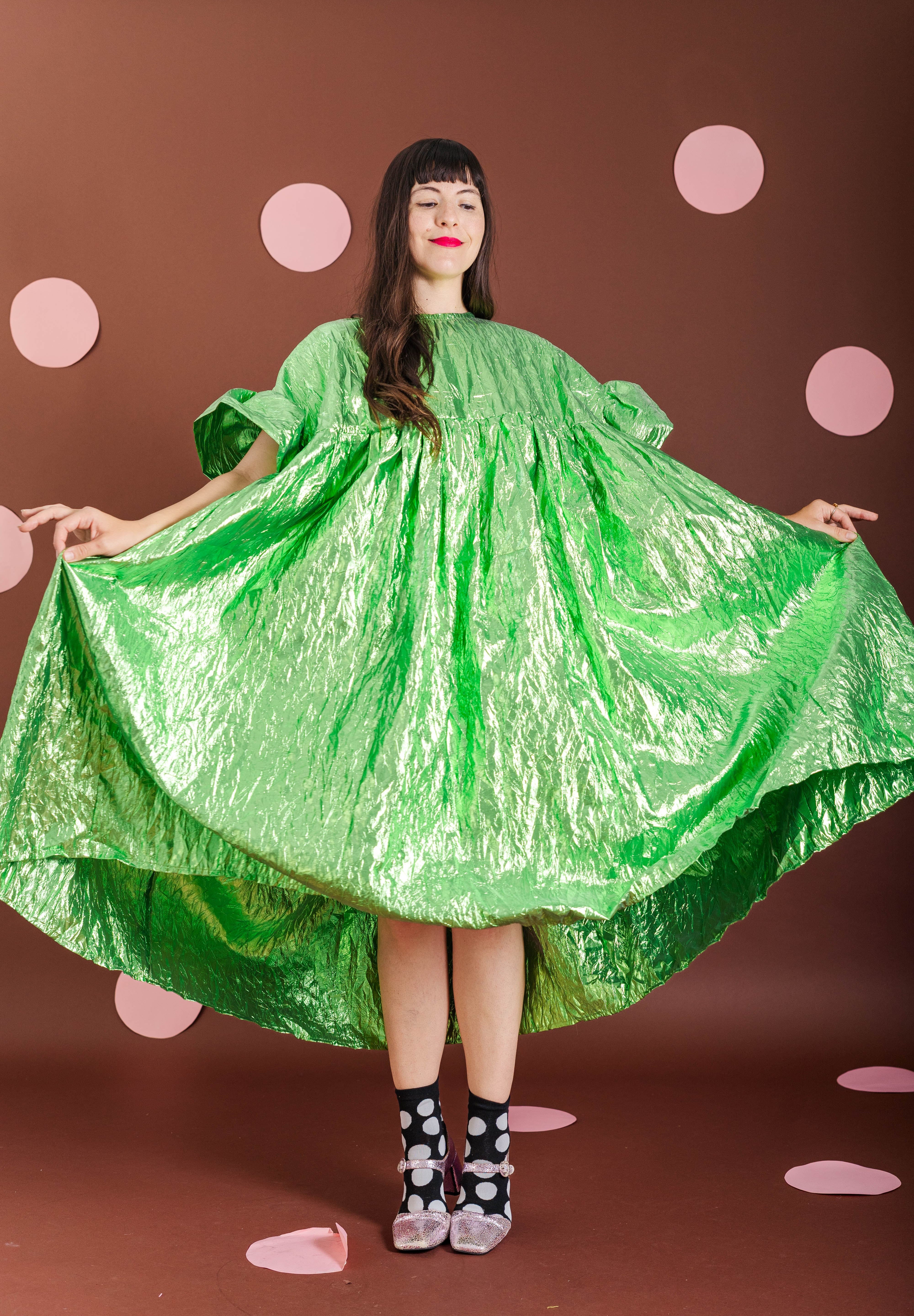 A woman wearing a voluminous, shiny bright green dress with short puff sleeves and a high-low hem, holding the dress out to show its shape against a brown backdrop with pink polka dots.