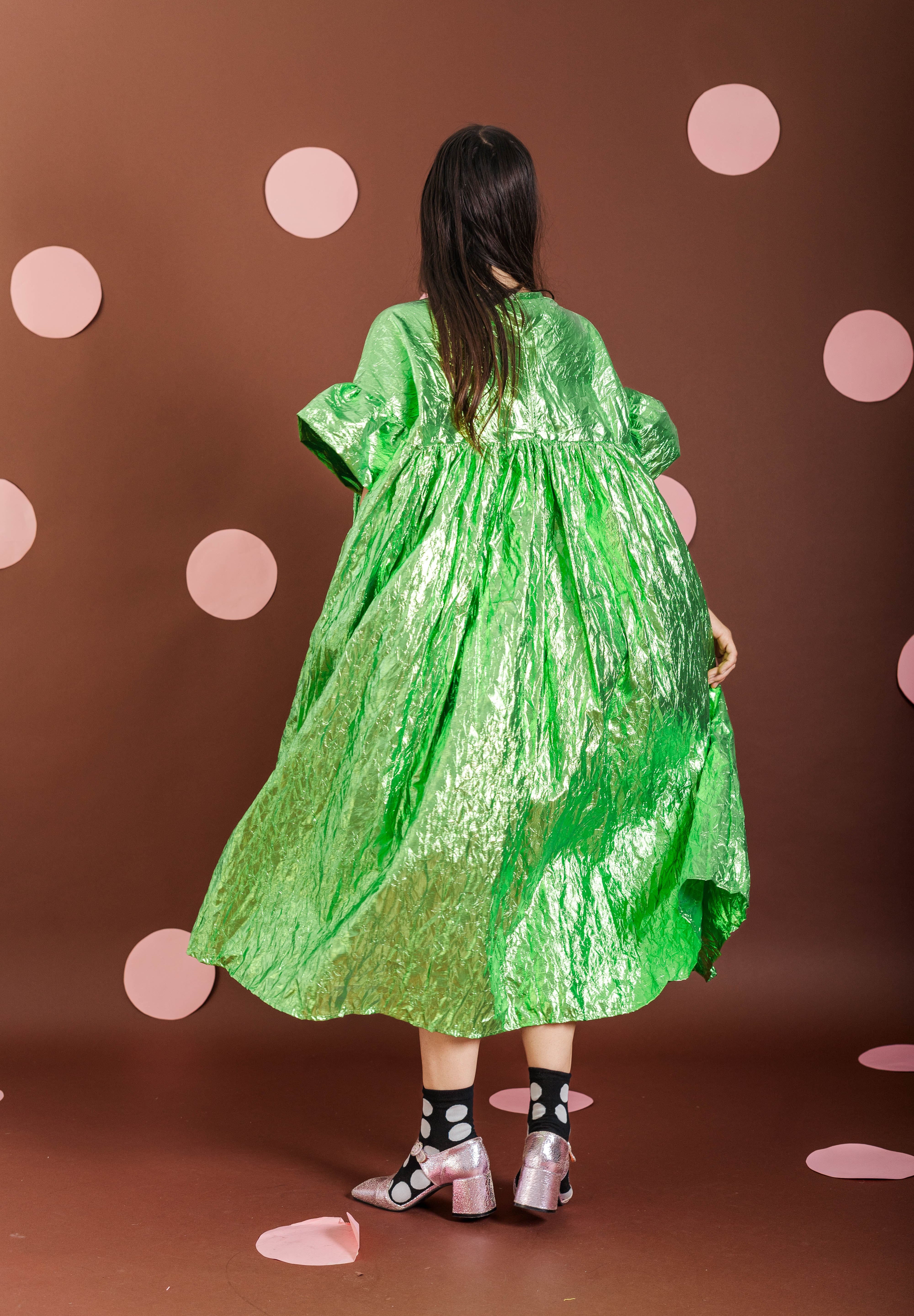 A model viewed from behind wearing a voluminous, shiny lime green dress with short puff sleeves, standing against a brown backdrop decorated with pink polka dots.