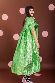 A woman modeling a voluminous, crinkled lime green dress with short wide sleeves, standing against a dark brown background decorated with pink polka dots.