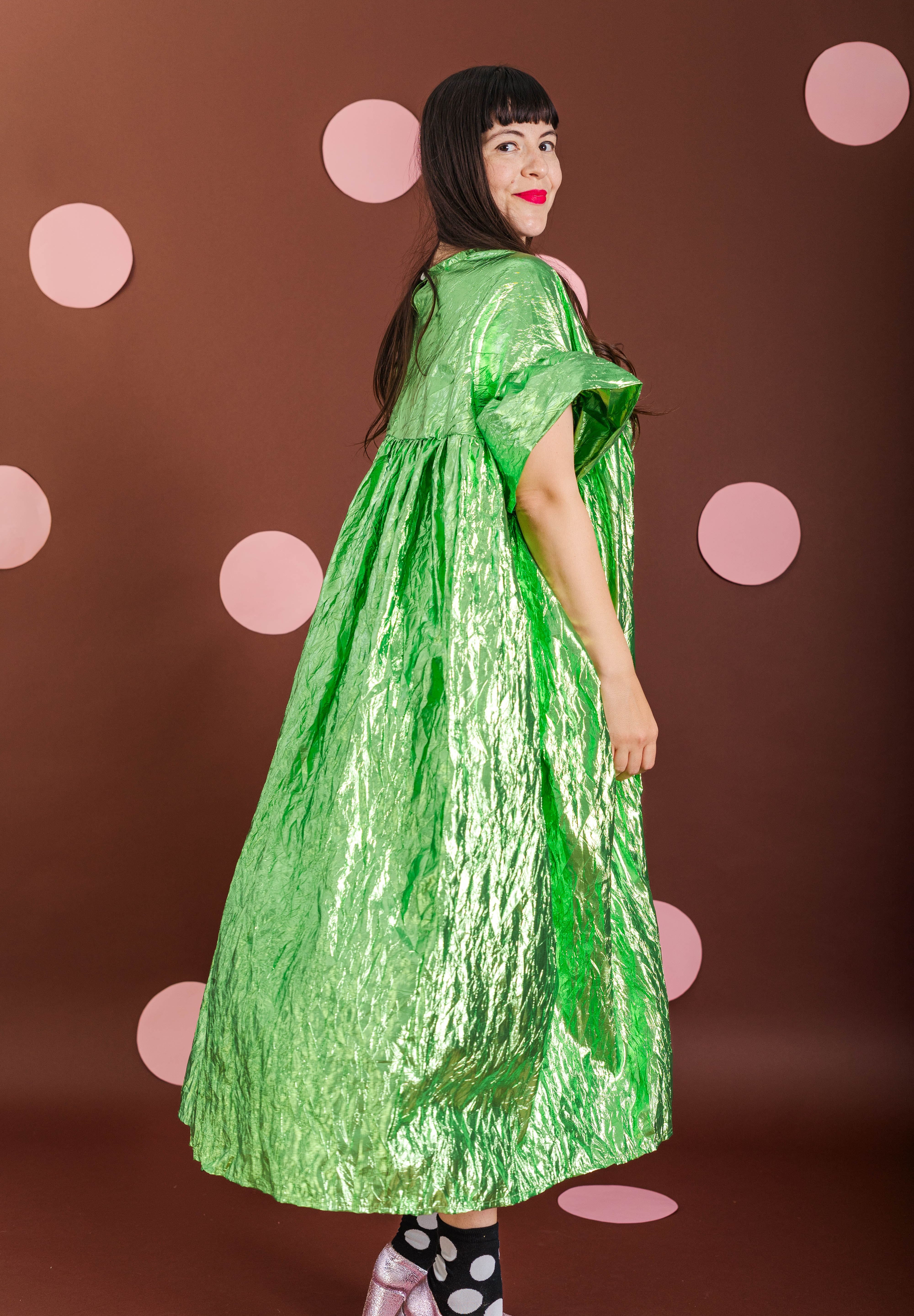 A woman modeling a voluminous, crinkled lime green dress with short wide sleeves, standing against a dark brown background decorated with pink polka dots.