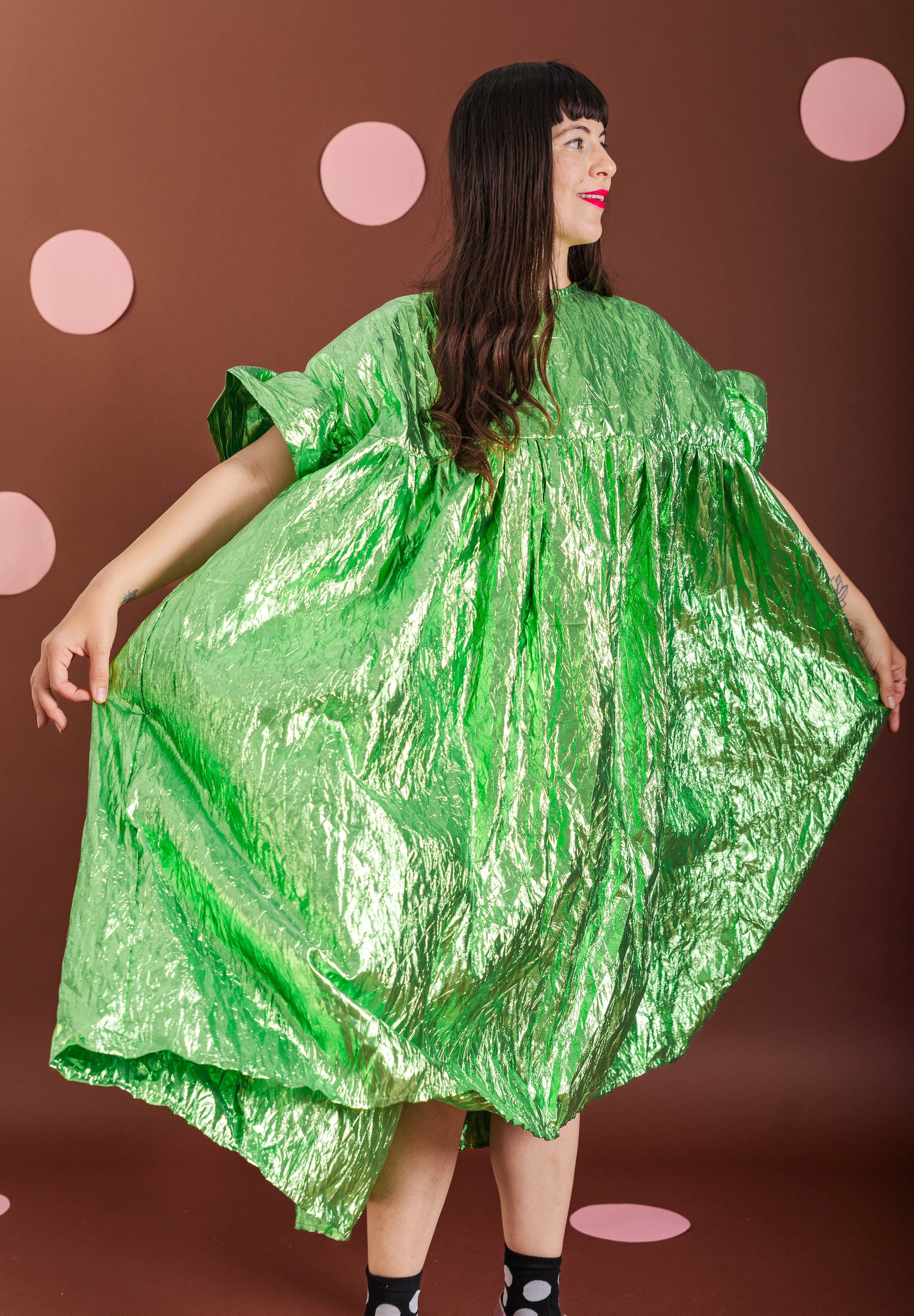 A woman modeling a voluminous, crinkled metallic green dress against a brown backdrop with pink polka dots.