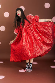 A woman wearing a voluminous, shiny red dress with puff sleeves twirls while holding out the skirt. She is wearing black and white polka-dot socks and standing against a brown backdrop decorated with pink polka dots.