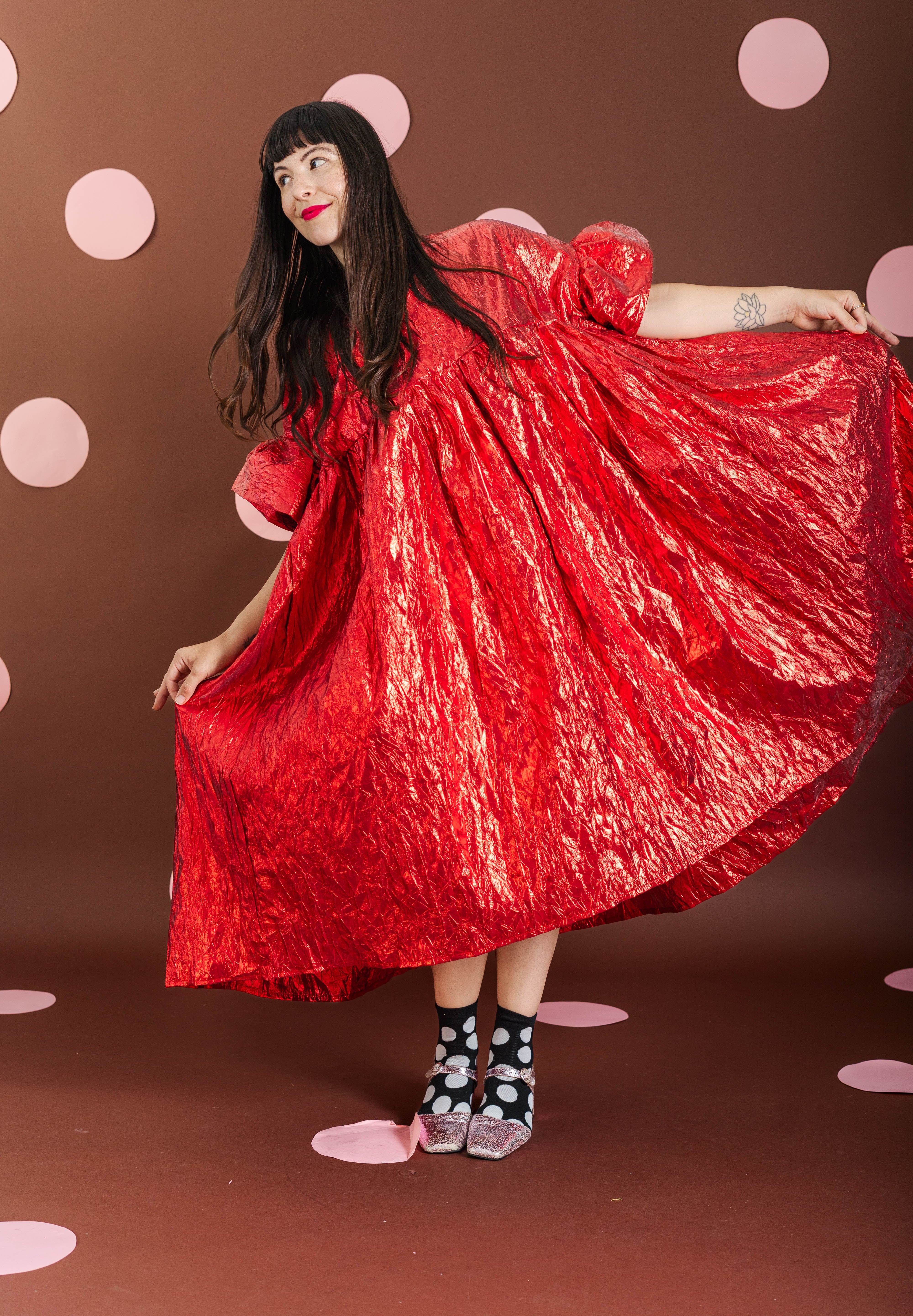 A woman wearing a voluminous, shiny red dress with puff sleeves twirls while holding out the skirt. She is wearing black and white polka-dot socks and standing against a brown backdrop decorated with pink polka dots.