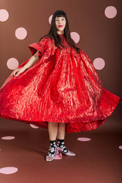 A woman wearing a voluminous, shiny red dress with short wide sleeves and a high gathered neckline, standing against a brown background with pink polka dots.