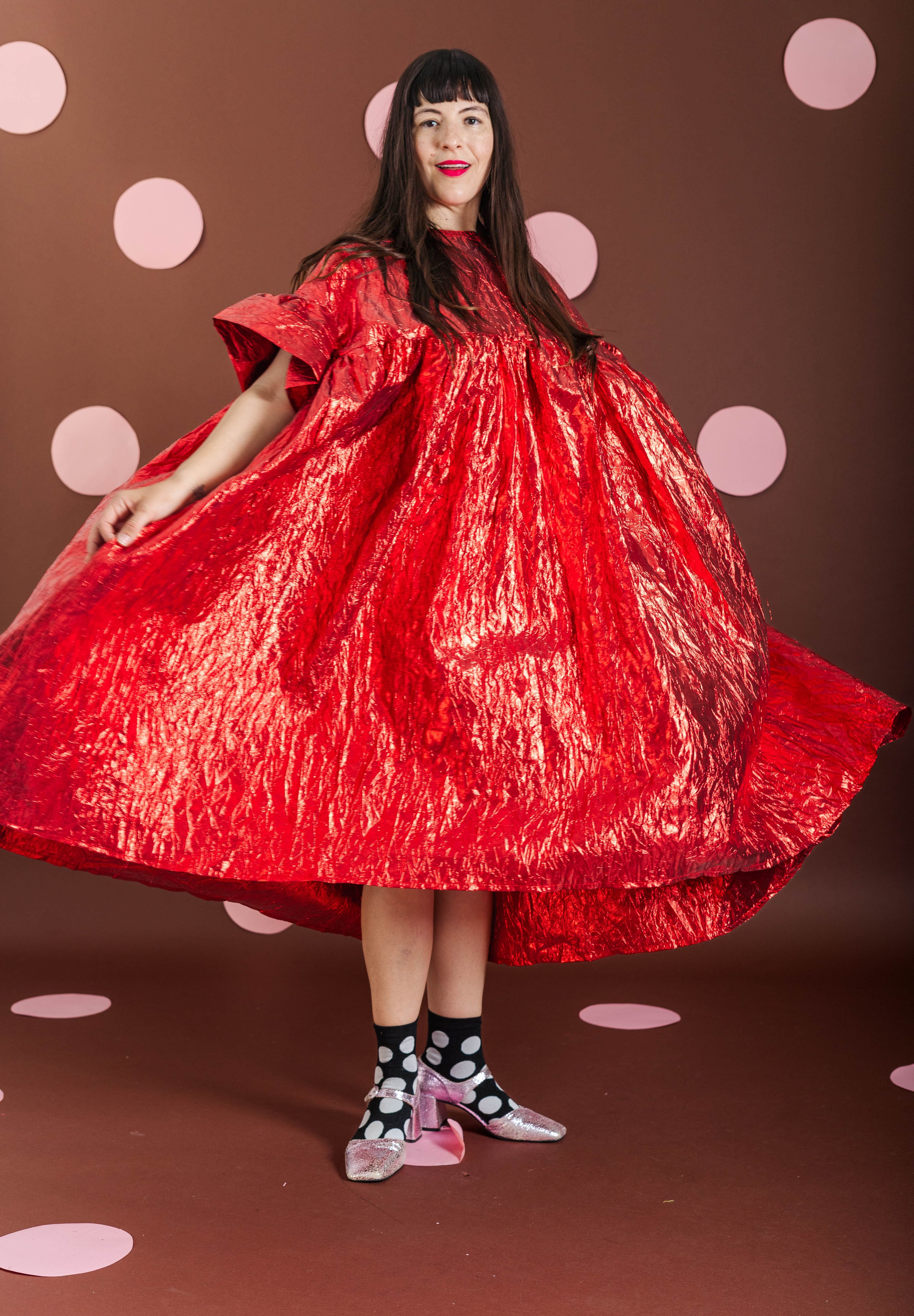 A woman wearing a voluminous, shiny red dress with short wide sleeves and a high gathered neckline, standing against a brown background with pink polka dots.