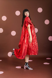 A woman posing in a shimmering red cape-style dress against a dark brown backdrop decorated with pink polka dots.