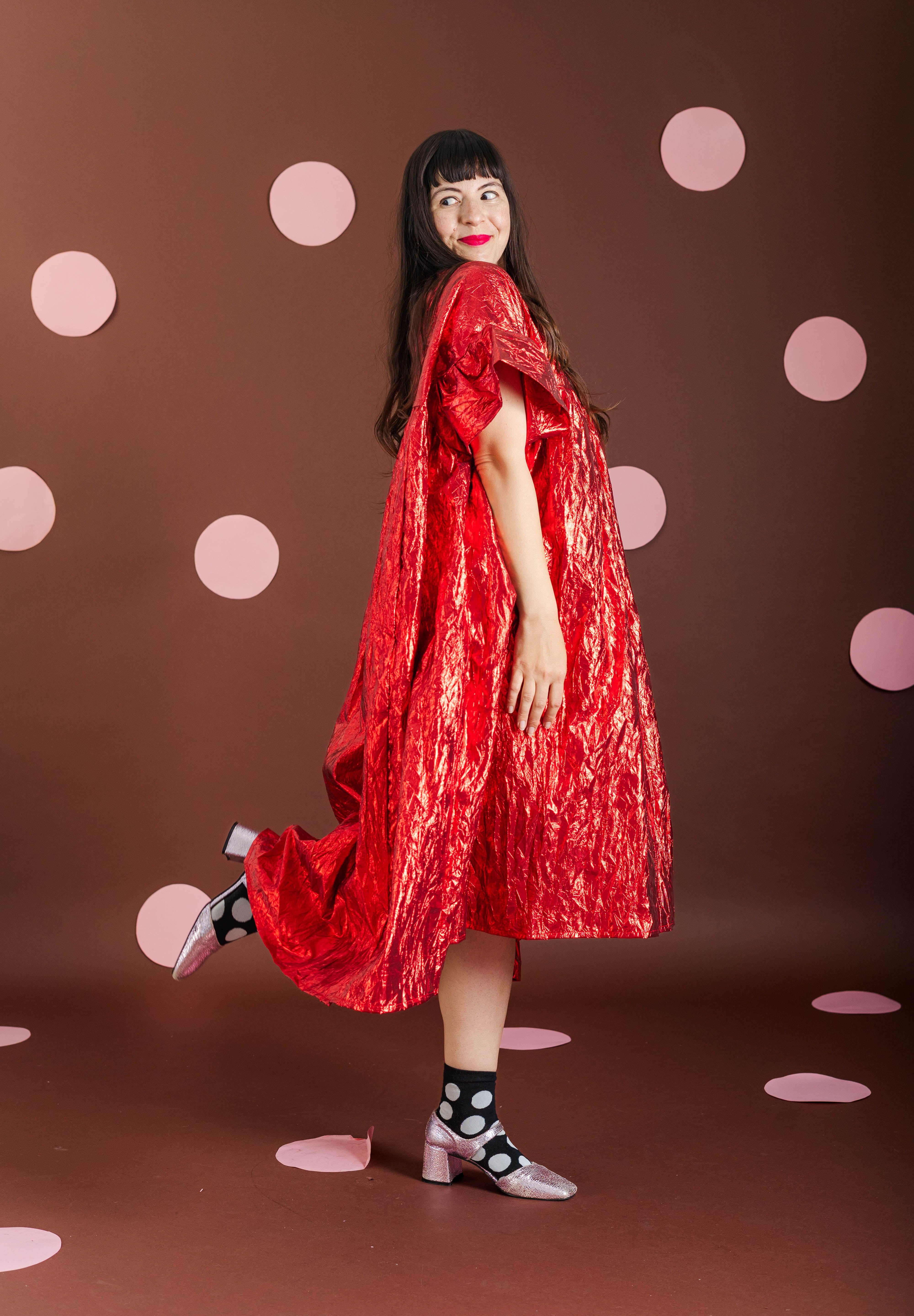 A woman posing in a shimmering red cape-style dress against a dark brown backdrop decorated with pink polka dots.