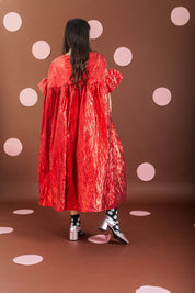 A model viewed from behind wearing a voluminous, crinkled red cape-style dress with puffed sleeves, standing against a brown backdrop decorated with light pink polka dots.