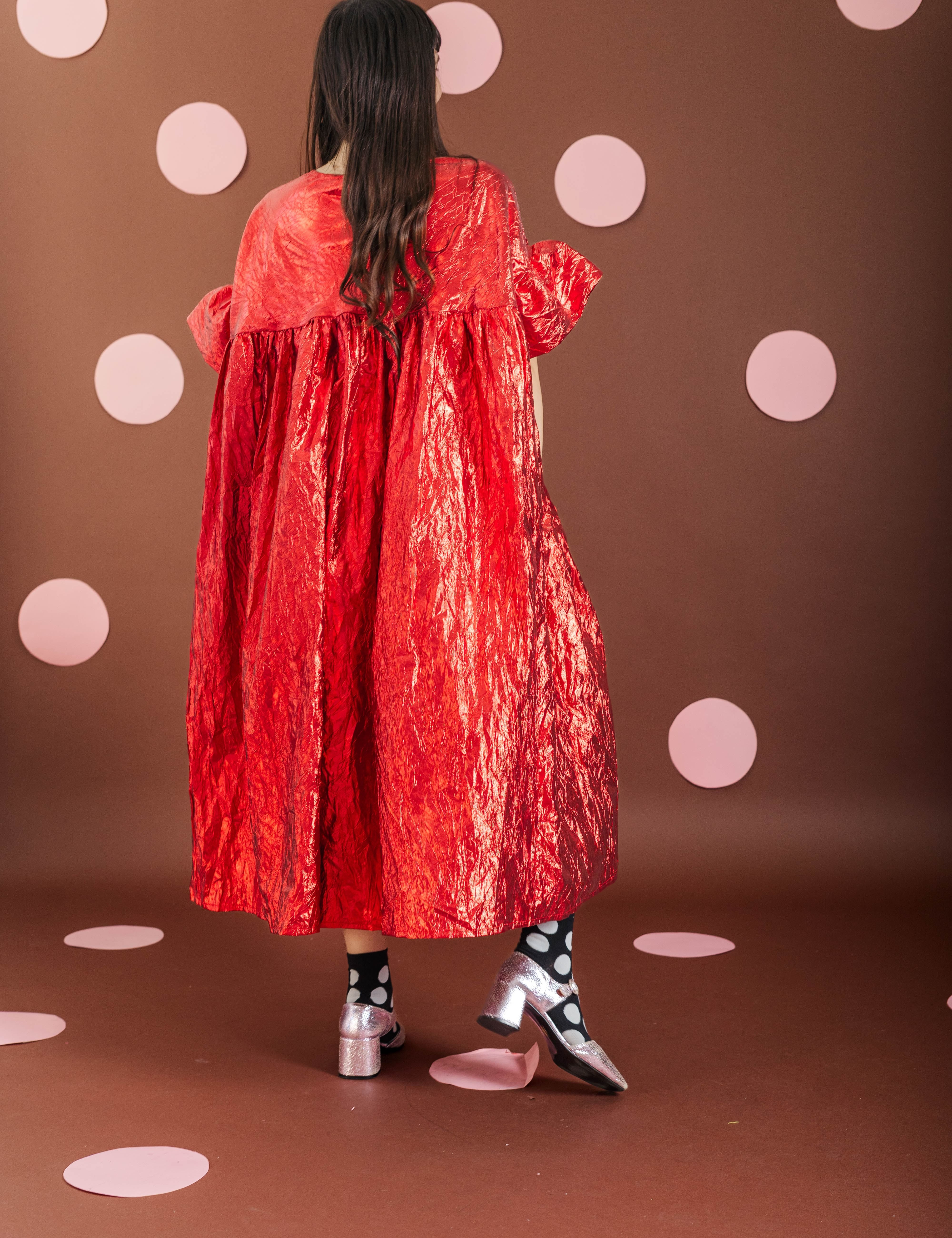 A model viewed from behind wearing a voluminous, crinkled red cape-style dress with puffed sleeves, standing against a brown backdrop decorated with light pink polka dots.