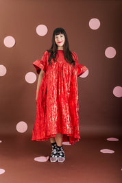 A woman modeling a shiny red high-low dress with wide short sleeves, standing against a brown backdrop decorated with pink polka dots.