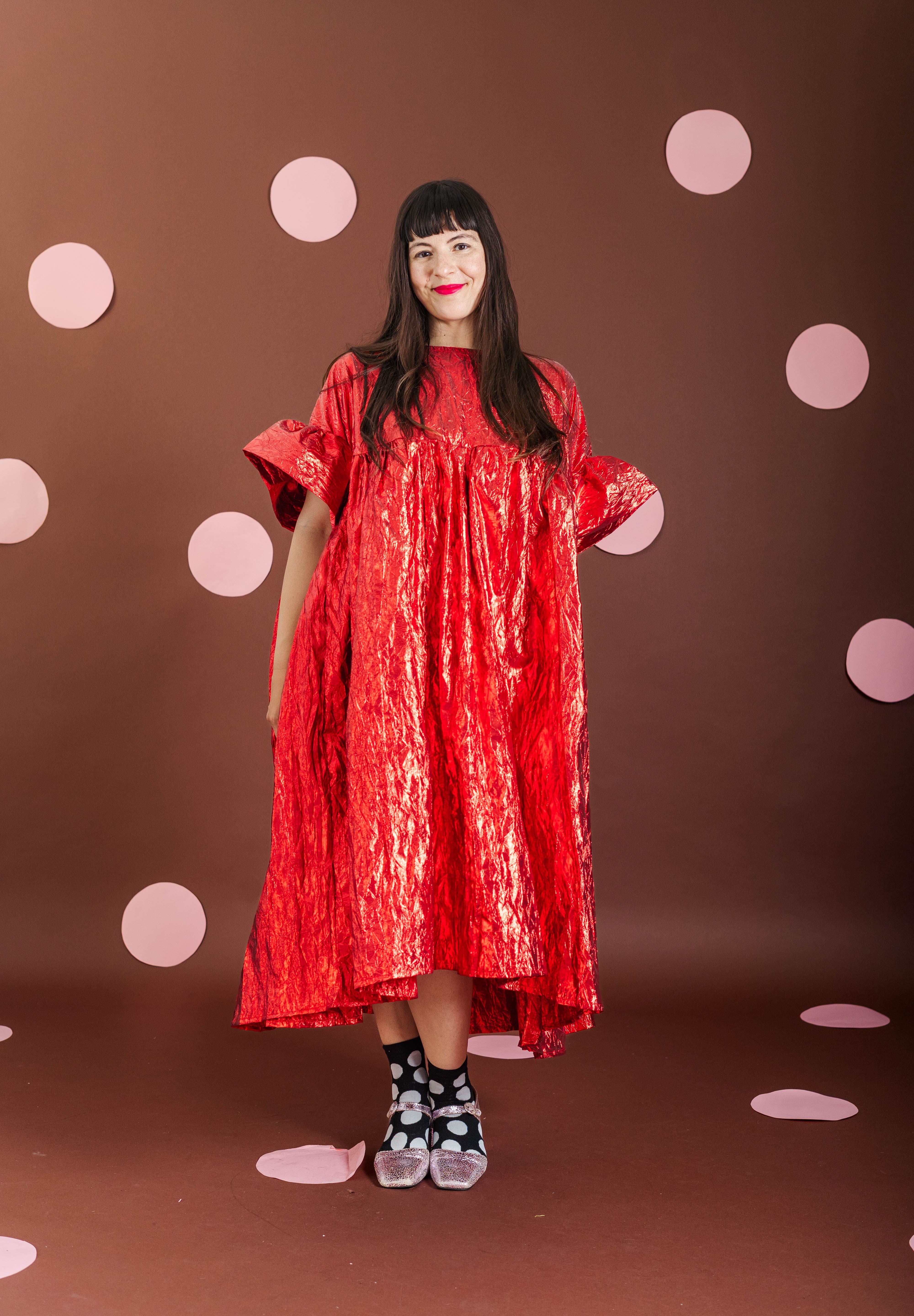 A woman modeling a shiny red high-low dress with wide short sleeves, standing against a brown backdrop decorated with pink polka dots.