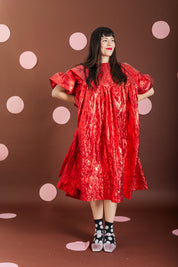A woman modeling a shiny red dress with voluminous short sleeves and a gathered silhouette, standing against a brown backdrop with pink polka dots.