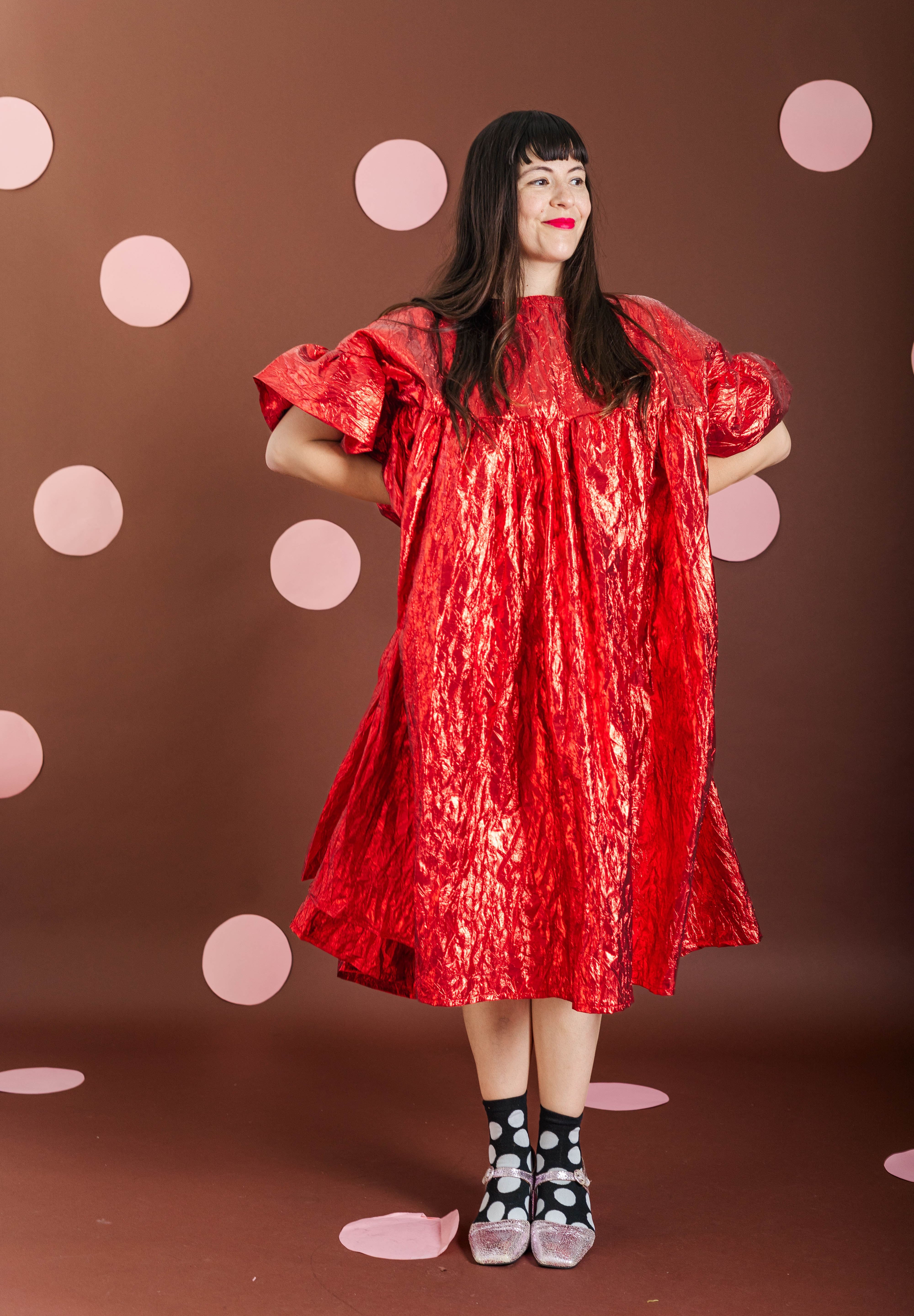 A woman modeling a shiny red dress with voluminous short sleeves and a gathered silhouette, standing against a brown backdrop with pink polka dots.